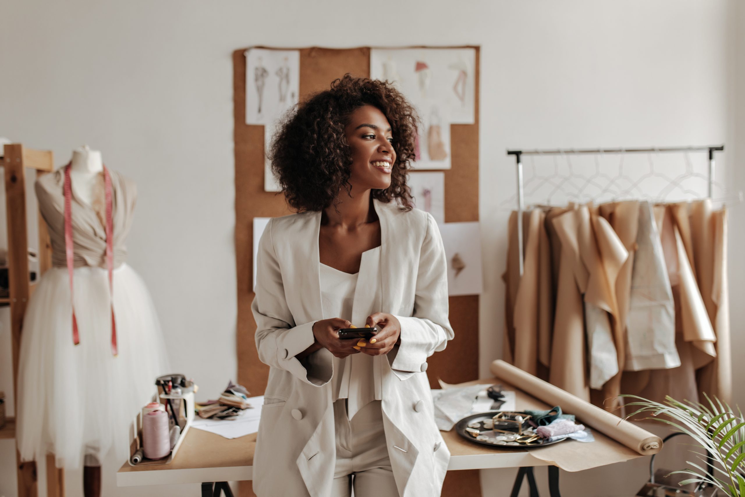 attractive-dark-skinned-curly-brunette-woman-oversized-jacket-white-pants-smiles-holds-phone-leans-table-office-fashion-designer-scaled